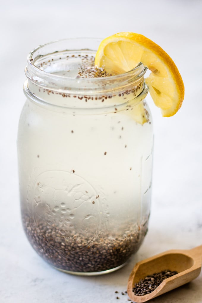 A large glass of water with chia seeds floating and on the bottom of the glass. There is a slice of lemon on the side and a small spoon full of chia seeds on the side