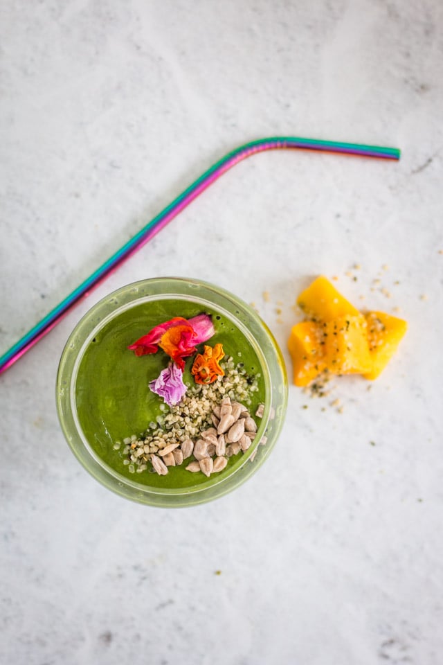 Mango avocado smoothie served in a glass with a multi-colored straw and topped with seeds and edible flowers