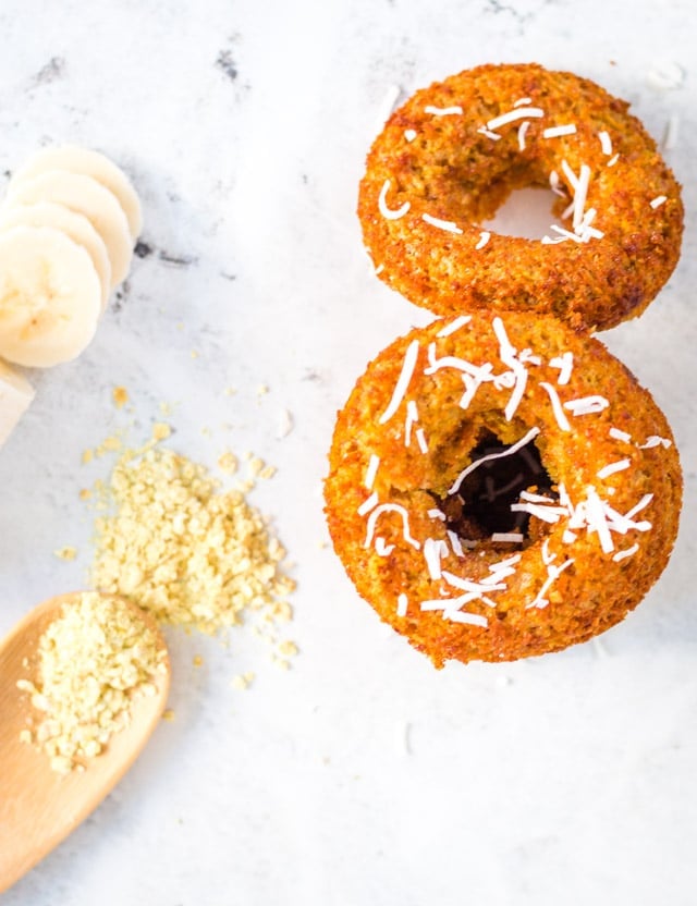 2 banana donuts on a white background with a wooden spoon holding raw oats and some banana slices in the background