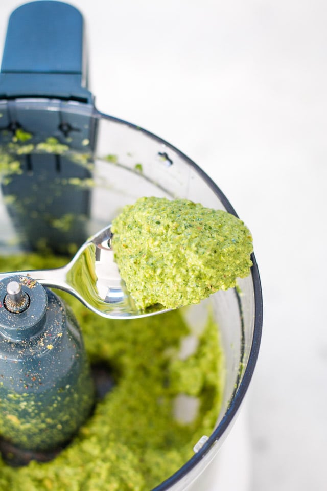 A spoon coming out of a food processor containing a light green carrot top pesto