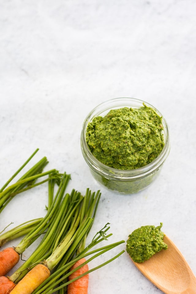 White background, jar of green carrot top pesto, bunch of colorful carrots with their tops removed and a small wooden spoon containing a mound of pesto