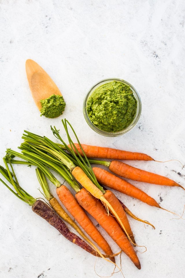 A bunch of colourful carrots with a jar of carrot top pesto and a wooden spoon with some pesto on it against a white background