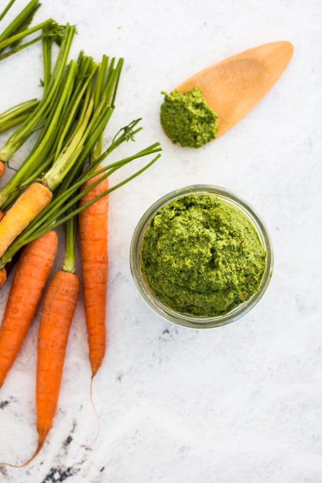 A bunch of colorful carrots with their tops removed, a glass jar of carrot top pesto and a small wooden spoon with a mound of pesto against a white backdrop.