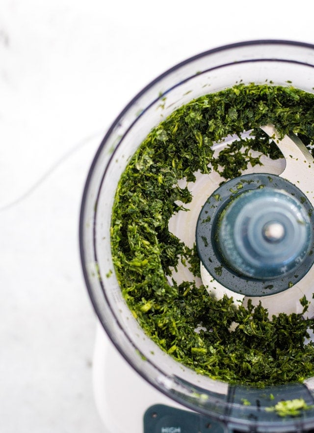 A food processor with green carrot top leaves and cilantro leaves pulsed until reduced in size