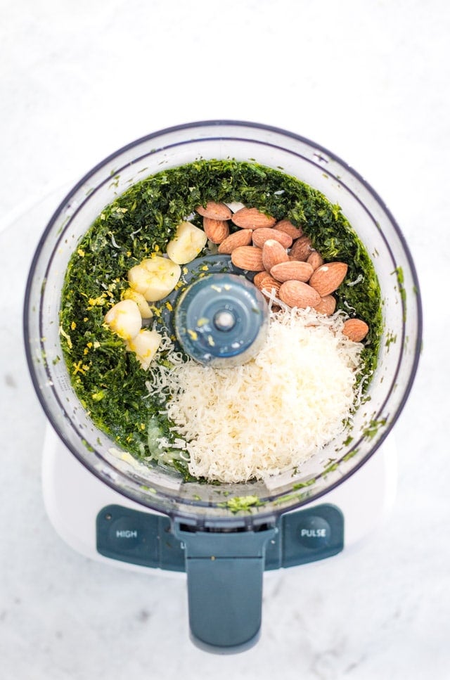 A food processor containing blitzed greens and topped with almonds, garlic, lemon and parmesan