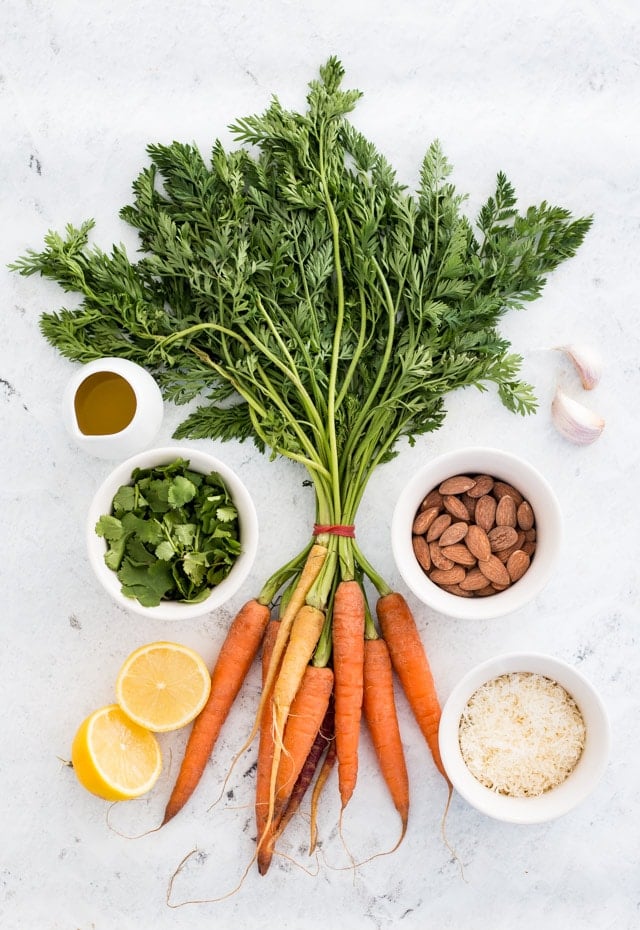 A bunch of carrots with tops still attached surrounded by lemons, cilantro, almonds, parmesan, garlic and olive oil on a white background.