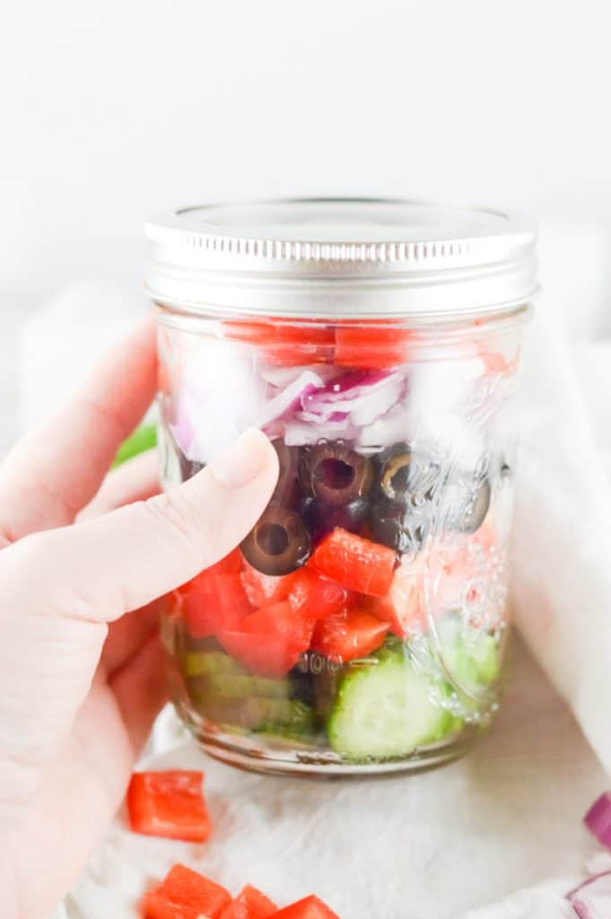 hand holding a glass mason jar with Greek salad ingredients inside