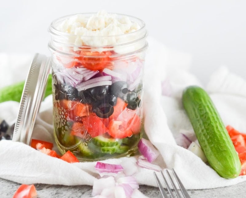 layered greek salad in a glass mason jar with cucumbers and tomatoes spread around in the white background