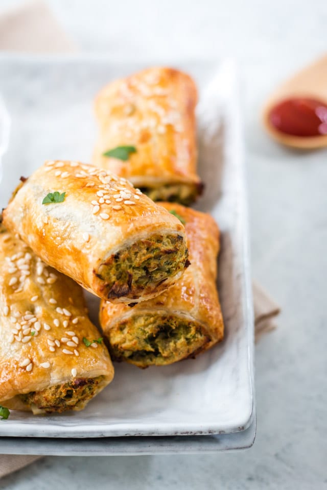 sausage rolls stacked on a white plate with a small wooden spoon containing a dollop of tomatoe ketchup in the background