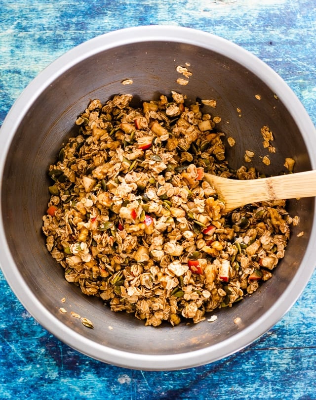 Combined ingredients for homemade apple cinnamon granola in a stainless steel bowl, being mixed with a wooden spoon.