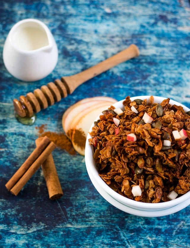 Homemade granola in a white bowl against a dark blue background with little pieces of fresh apple sprinkled on top and to the side