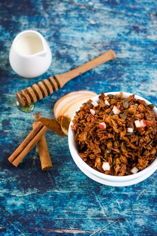 Apple Cinnamon Granola in a white bowl against a dark blue background with a little pot of milk, honey stick and cinnamon sticks