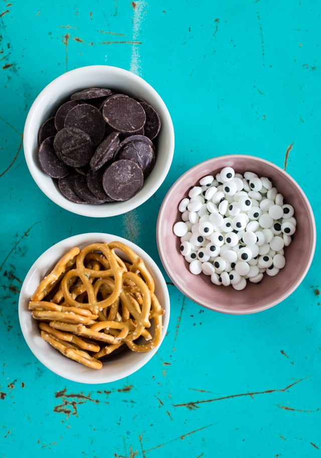 teal painted metal background with 3 bowls containing ingredients: pretzels, chocolate discs and google eyes