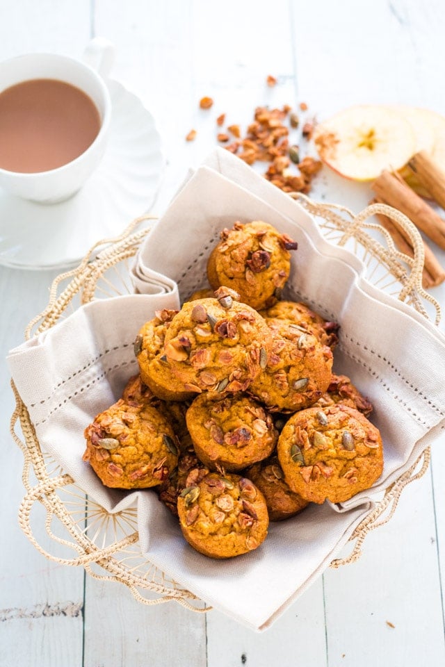 pumpkin muffins in a small basket lined with a linen napkin. apple slices and cinnamon sticks in the background, along with a cup of tea and saucer