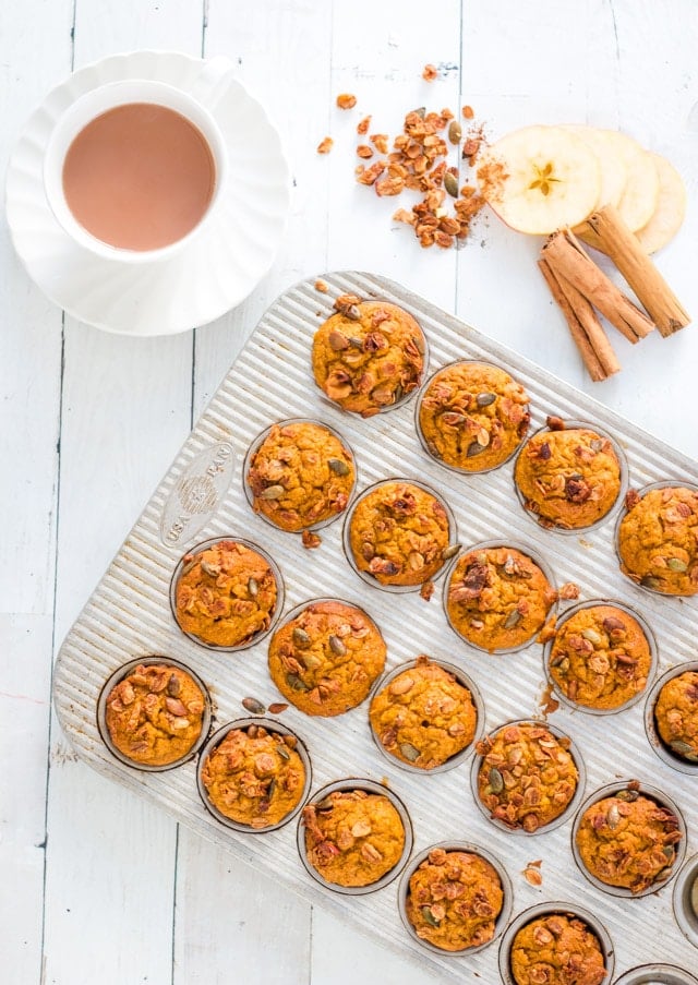 Mini muffin tray loaded with cooked mini pumpkin muffins. apple slices, cinnamon sticks and a sprinkling of granola against a white background.