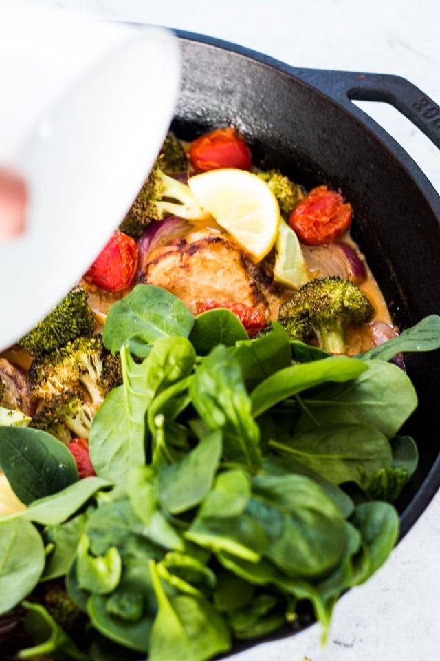 Baby spinach being added to a cast iron skillet containing chicken and mixed vegetables