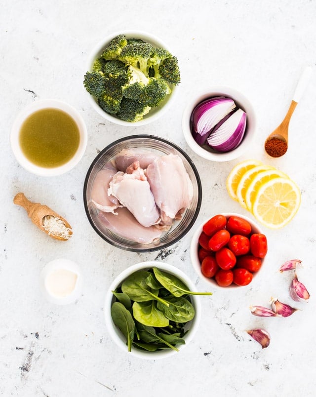 A white background with bowls containing chicken, tomatoes, broccoli, spinach, red onion, lemon, parmesan and garlic