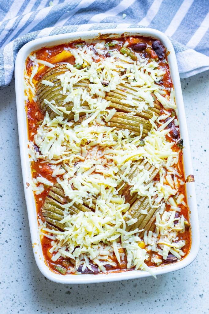 Cheese being sprinkled on top of chili and potatoes in a white casserole dish