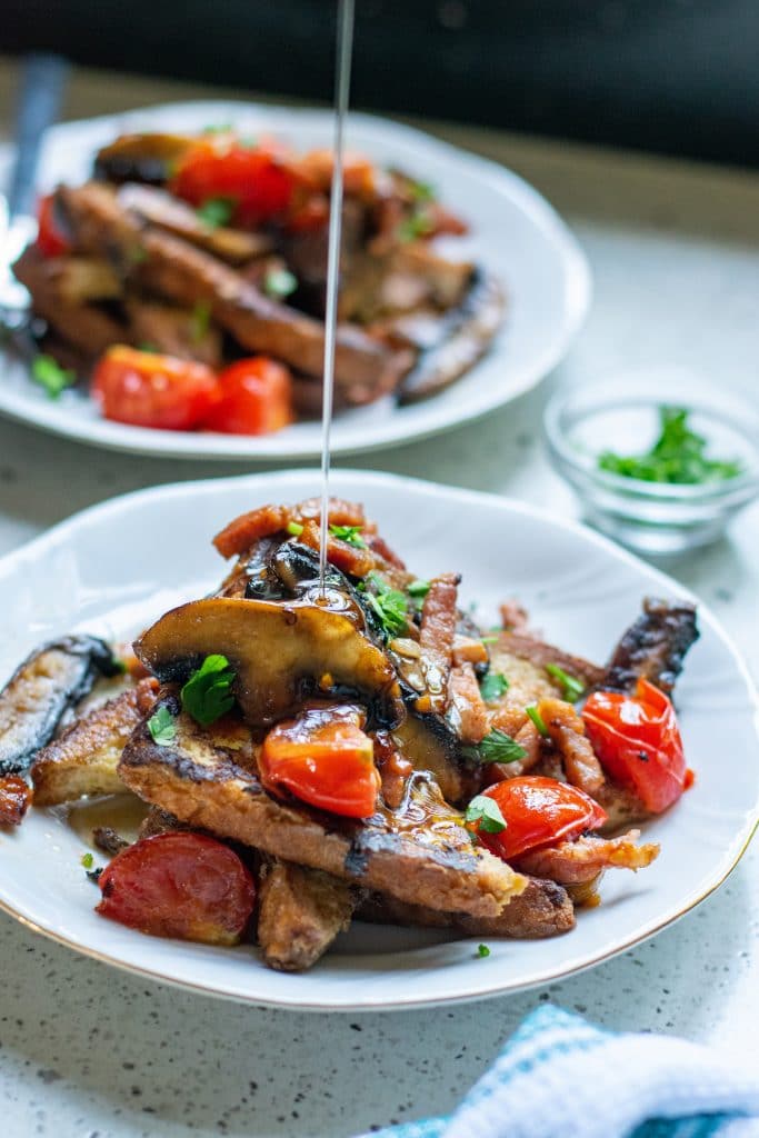 Maple syrup being drizzled on top of french toast stacked with mushrooms, bacon and cherry tomatoes