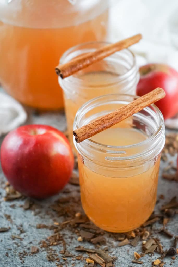 Two glasses of instant pot apple cider served in mason jars with cinnamon sticks on top and apples in the background