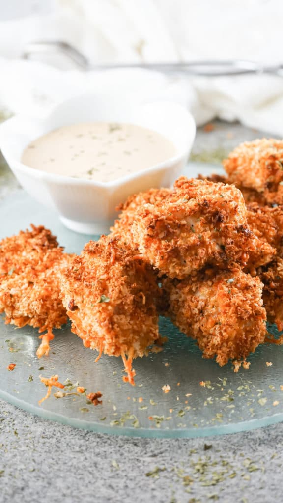 Air Fryer Chicken Nuggets piled onto a glass serving dish with a white bowl in the background containing a dipping sauce