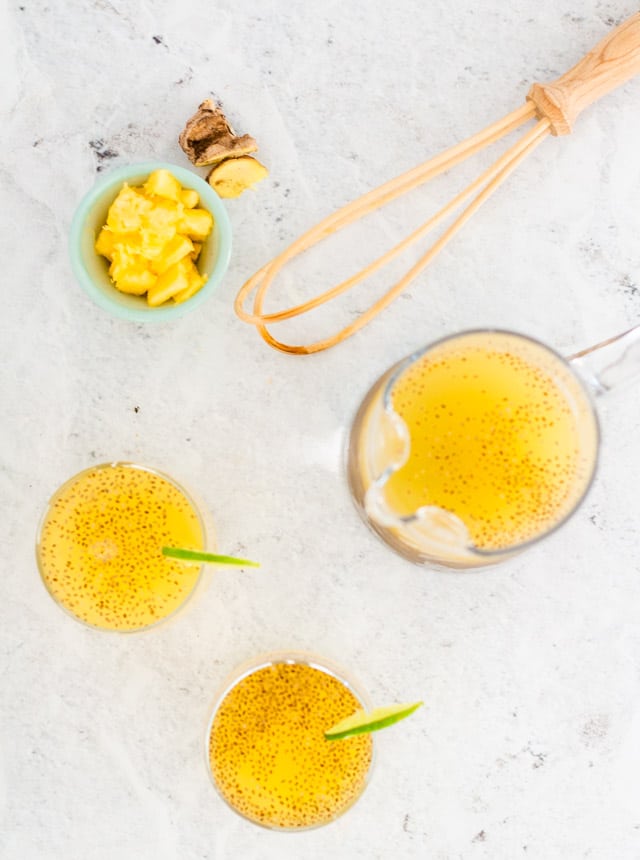 Overhead flat lay of pineapple agua fresca with a full jug, two smaller glasses, a whisk and some pineapple and ginger