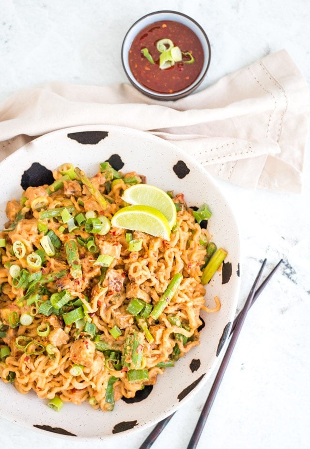 Spicy peanut noodles served in a black and white bowl against a white background with a small bowl of sweet chili sauce