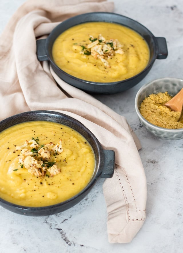 Overhead shot of cauliflower and leek soup served in two dark grey bowls with some nutritional yeast about to be scooped on top