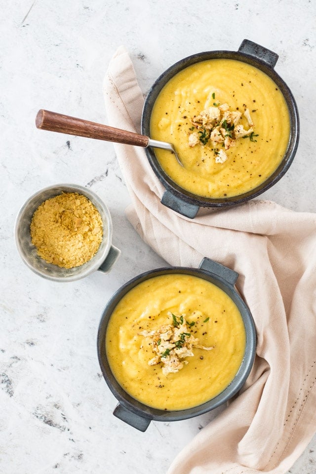 Overhead shot of cauliflower soup served into two dark grey bowls with a linen napkin and a bowl of nutritional yeast on the side