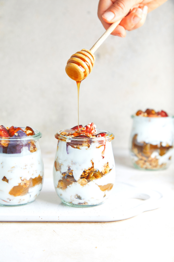 Layered fruit and yogurt parfaits in 3 small glass jars with honey being drizzled on top