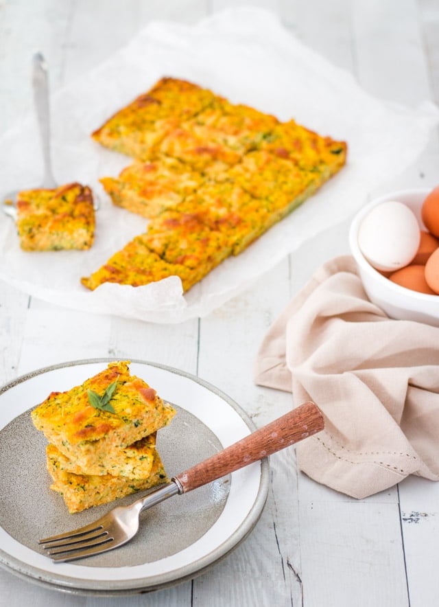 Grey plate with a stack of 3 slices of zucchini slice and a wooden fork ready to serve it up. The rest of the gluten free slice is in the background.