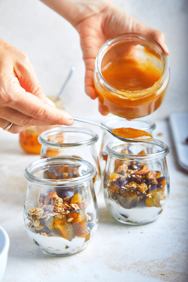 Fruit yogurt parfait being constructed: ingredients have been layered in two small glass jars and some honey is being drizzled on top