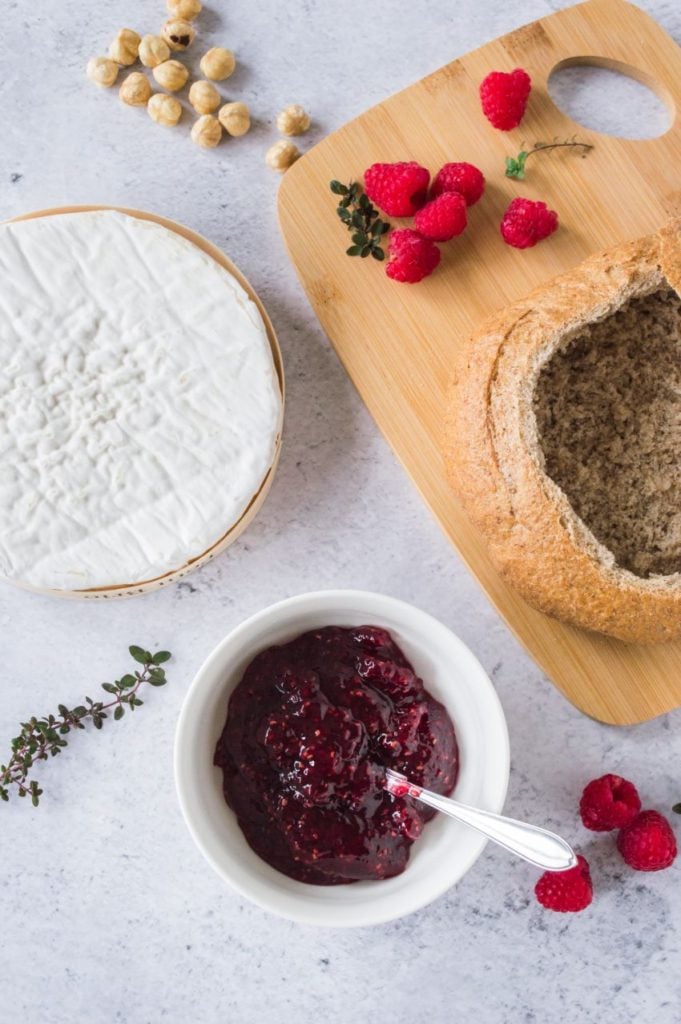 Overhead flatlay shot of ingredients required for a baked brie: hollowed out cob loaf, raspberry jam, brie round and some fresh raspberries and thyme