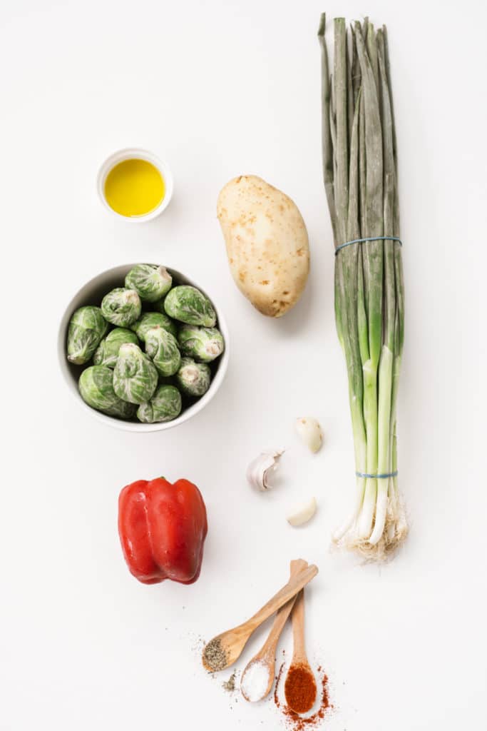 overhead flat lay ingredient shot showing potaotes, brussels sprouts, red peppers, garlic, spring onions and some spices