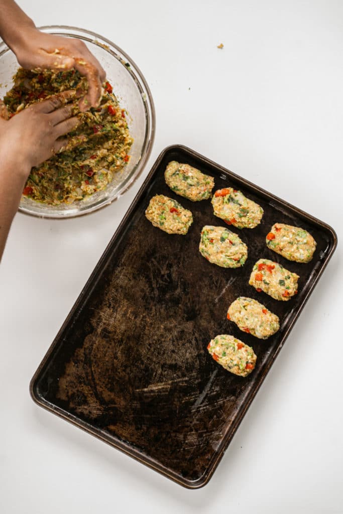 uncooked vegetable tots being line on an oiled baking tray with a hand getting more mixture from a glass bowl on the side