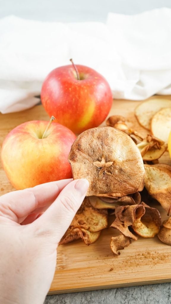 air fryer apple chips being picked up from a wooden board with fresh apples in the background