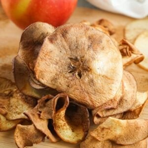 A stack of crispy apple chips on a wooden board with a fresh apple in the background