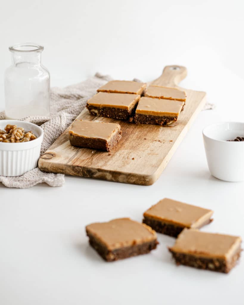 brownie slices on a wooden chopping board with bowls of nuts on the side against a white background.