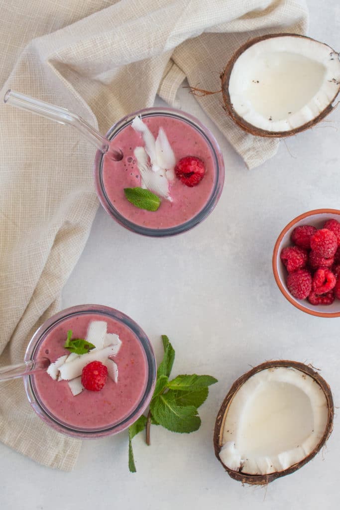 red berry protein smoothies served in 2 glasses with glass straws with fresh raspberries and coconut in the background