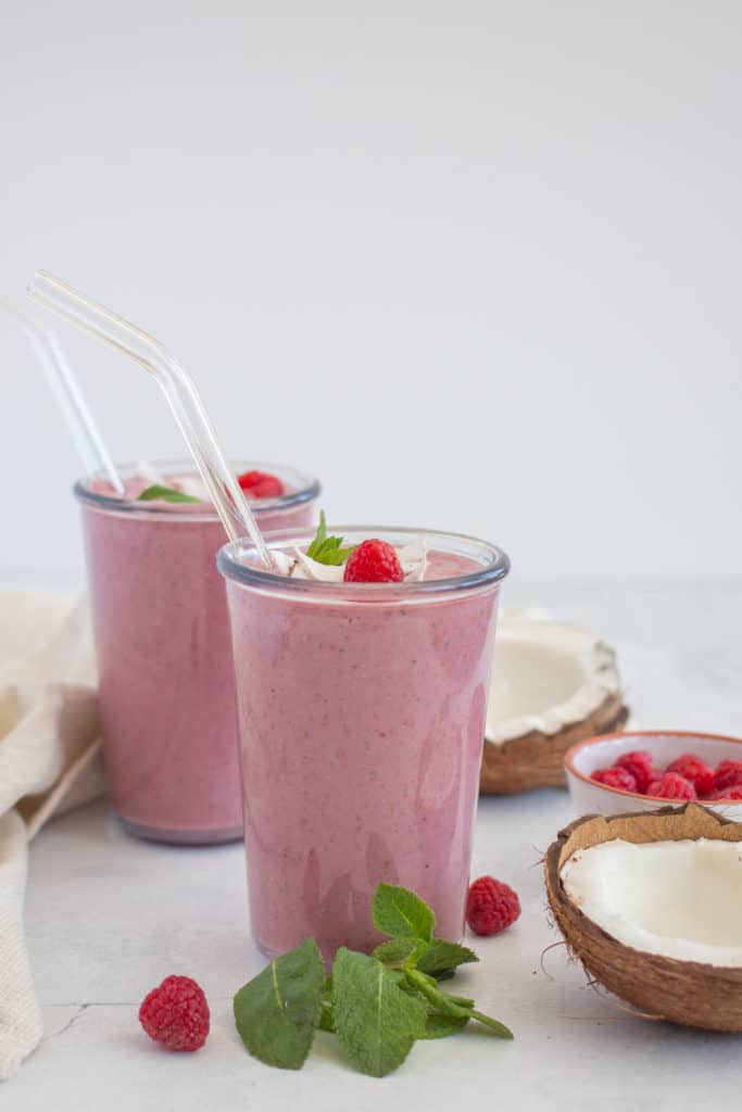 red berry protein smoothies served in 2 glasses with glass straws with fresh raspberries and coconut in the background