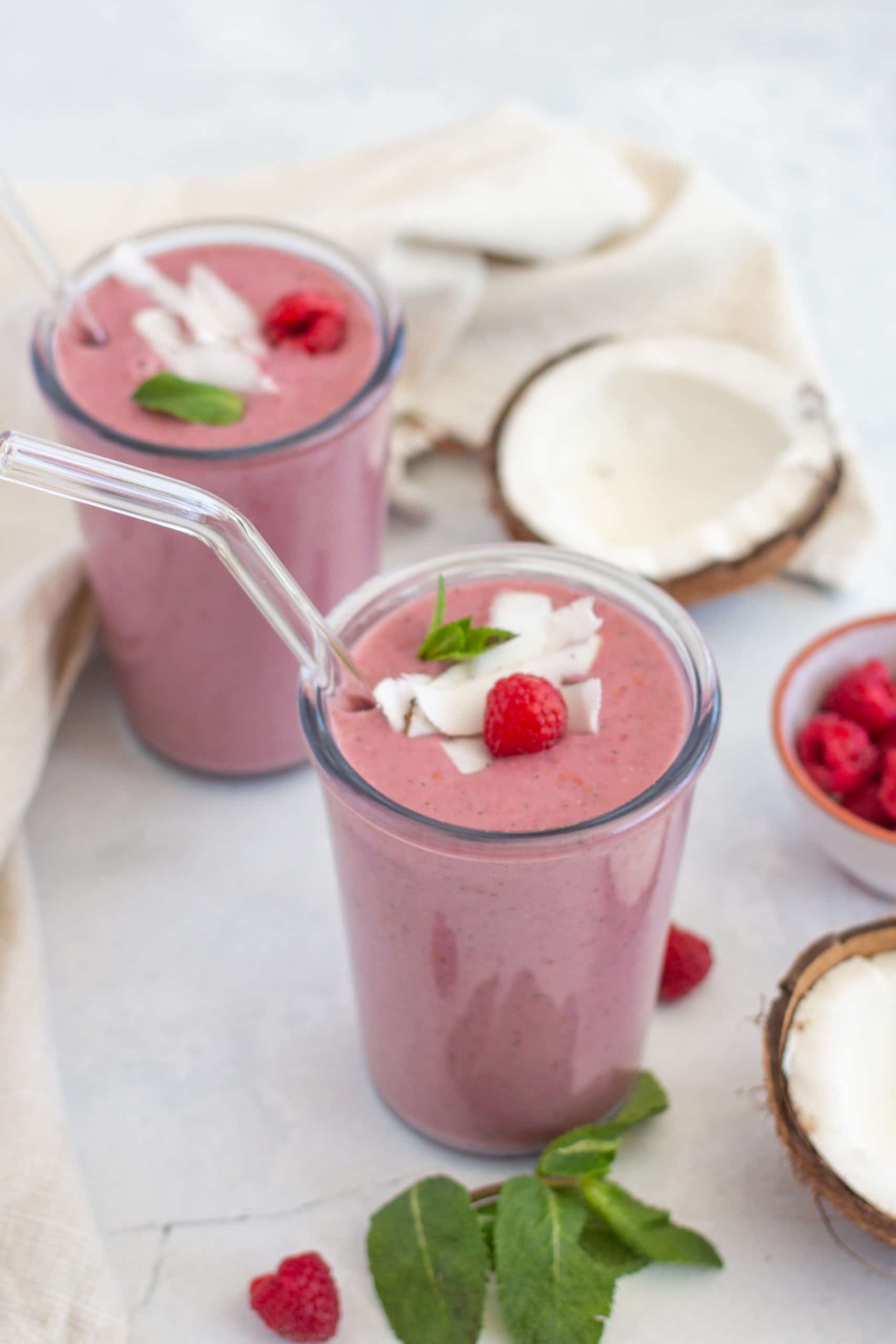 red berry protein smoothies served in 2 glasses with glass straws with fresh raspberries and coconut in the background