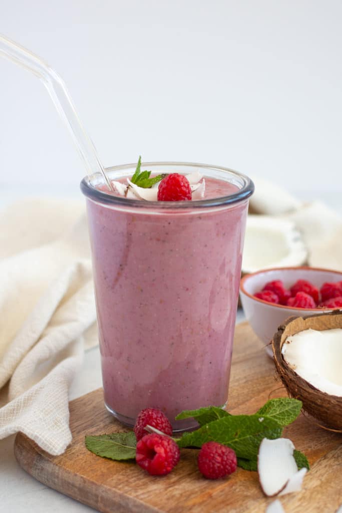 red berry smoothie in a tall glass placed on a chopping board and surrounded by berries and spinach