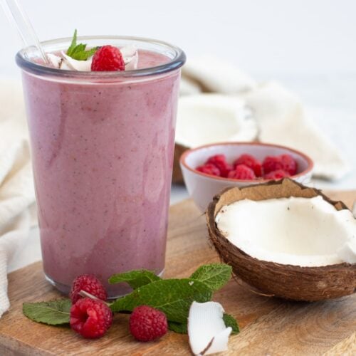 red berry smoothie in a tall glass placed on a chopping board and surrounded by berries and spinach