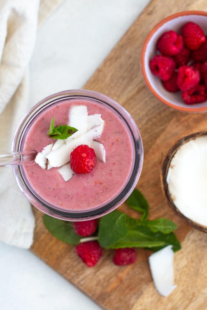 red berry smoothie in a tall glass placed on a chopping board and surrounded by berries and spinach