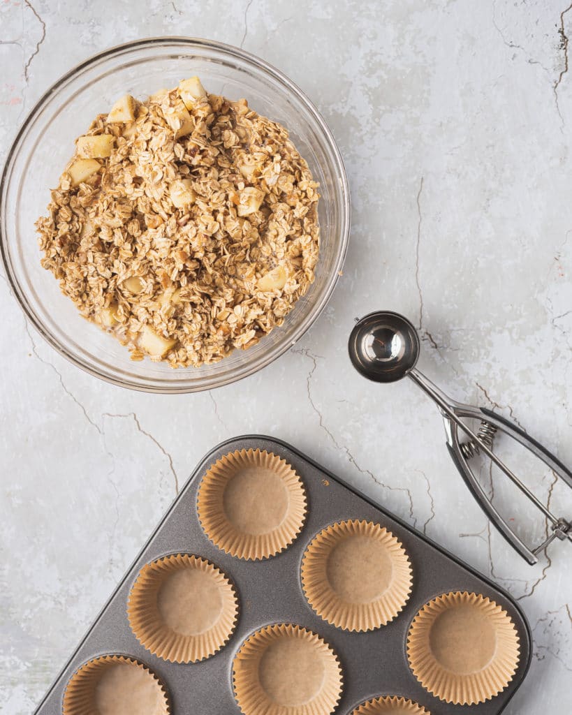 overhead shot of mixed apple cinnamon oatmeal cup ingredients with a cookie scoop and a muffin tray to the side