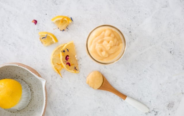 overhead shot of lemon curd in a glass jar with a sprinkling of lemon slices and edible flowers in the background