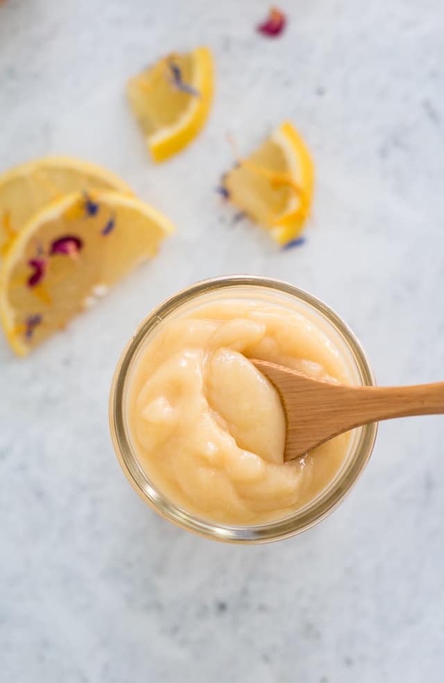 overhead shot of lemon curd in a glass jar with a sprinkling of lemon slices and edible flowers in the background