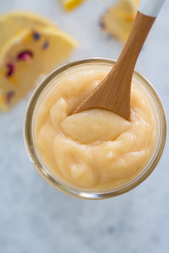 overhead shot of lemon curd in a glass jar with a sprinkling of lemon slices and edible flowers in the background