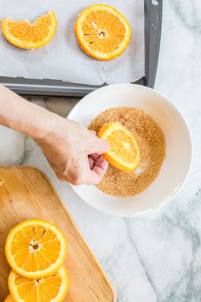 Overhead shot of a hand dipping orange slices into turbinado sugar and then laying them on a baking sheet lined with parchment paper