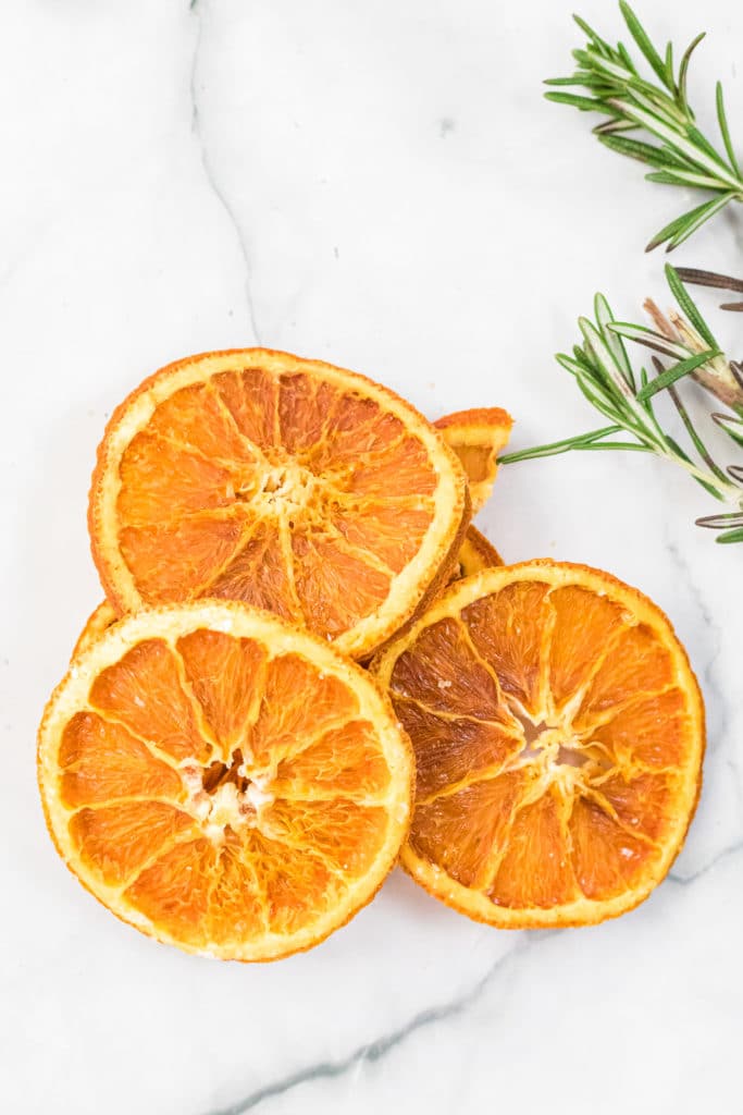 Dried orange slices stacked together against a white background, with some rosemary sprigs in the background.